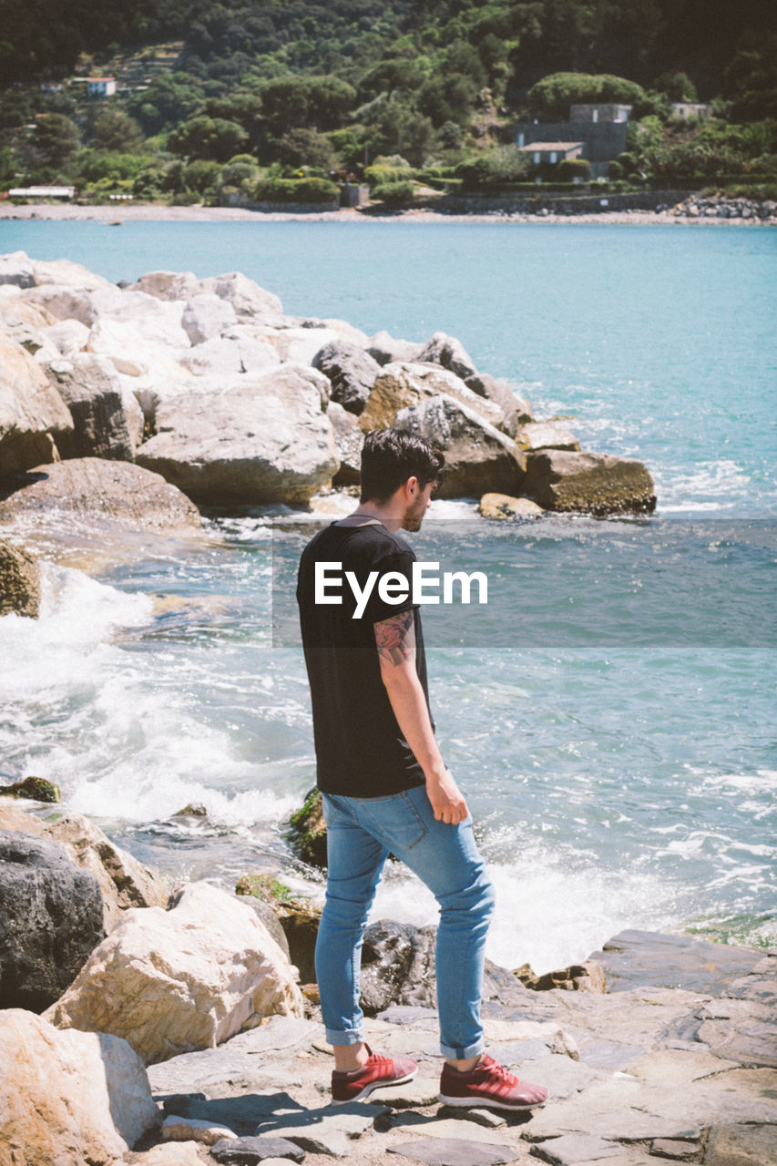 REAR VIEW OF YOUNG MAN STANDING ON ROCK AT SEA SHORE