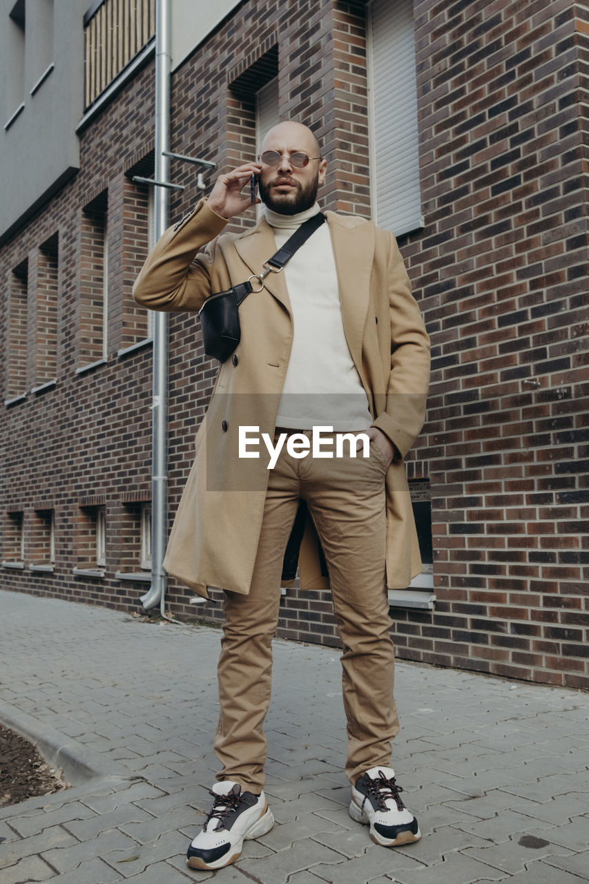 Portrait of man talking on phone while standing against building on footpath