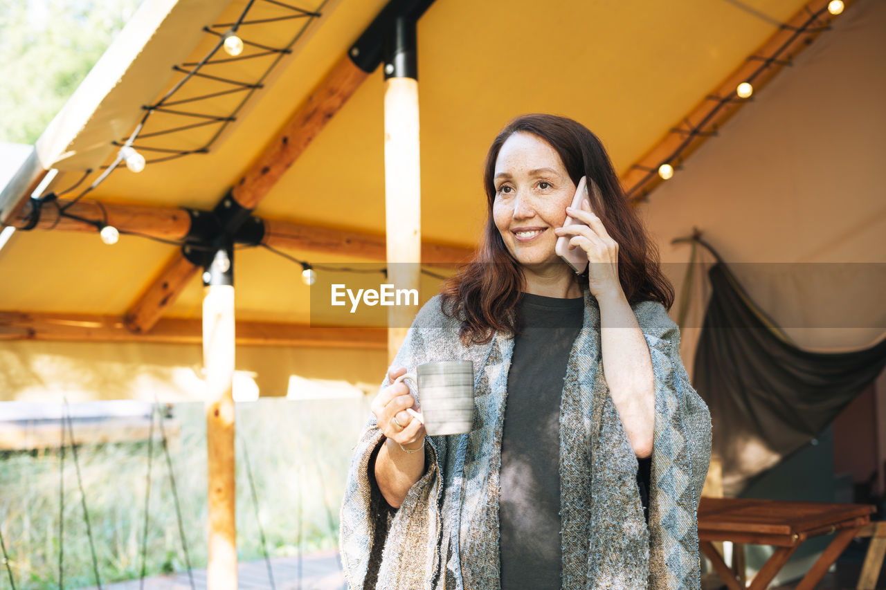 Brunette woman in poncho using mobile phone and relaxing in glamping in nature