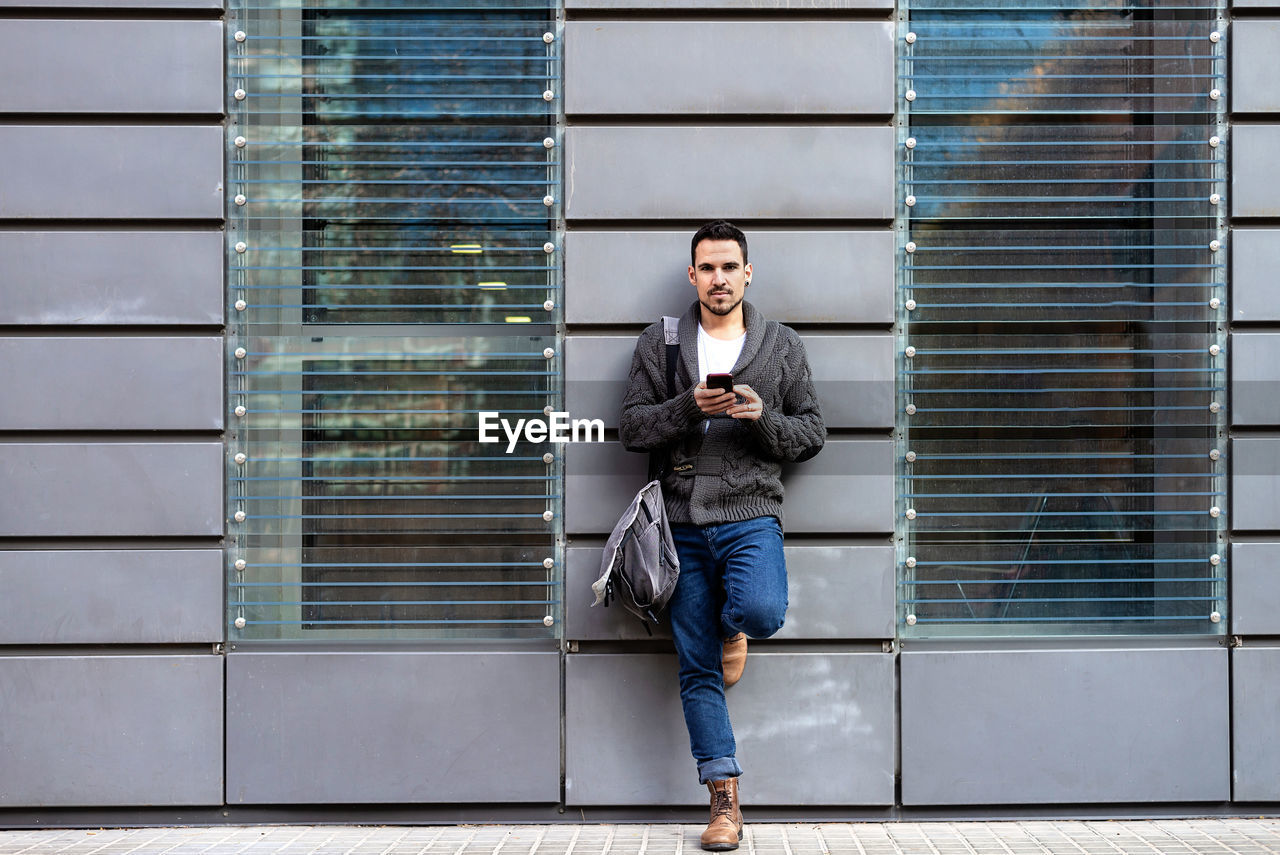 Front view of a bearded man using phone leaning on office building wall