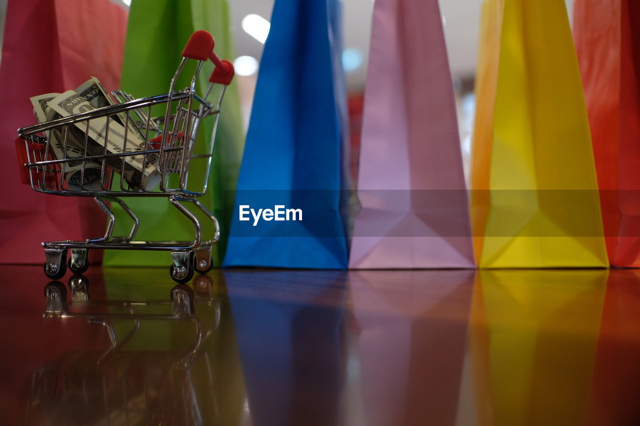 Close-up of colorful shopping bags with paper currencies in miniature shopping cart on wooden table
