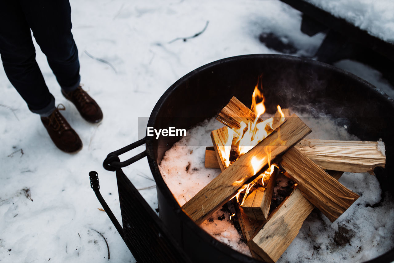 High angle of crop unrecognizable traveler standing on snow and warming up near camping fireplace with burning firewood in winter day