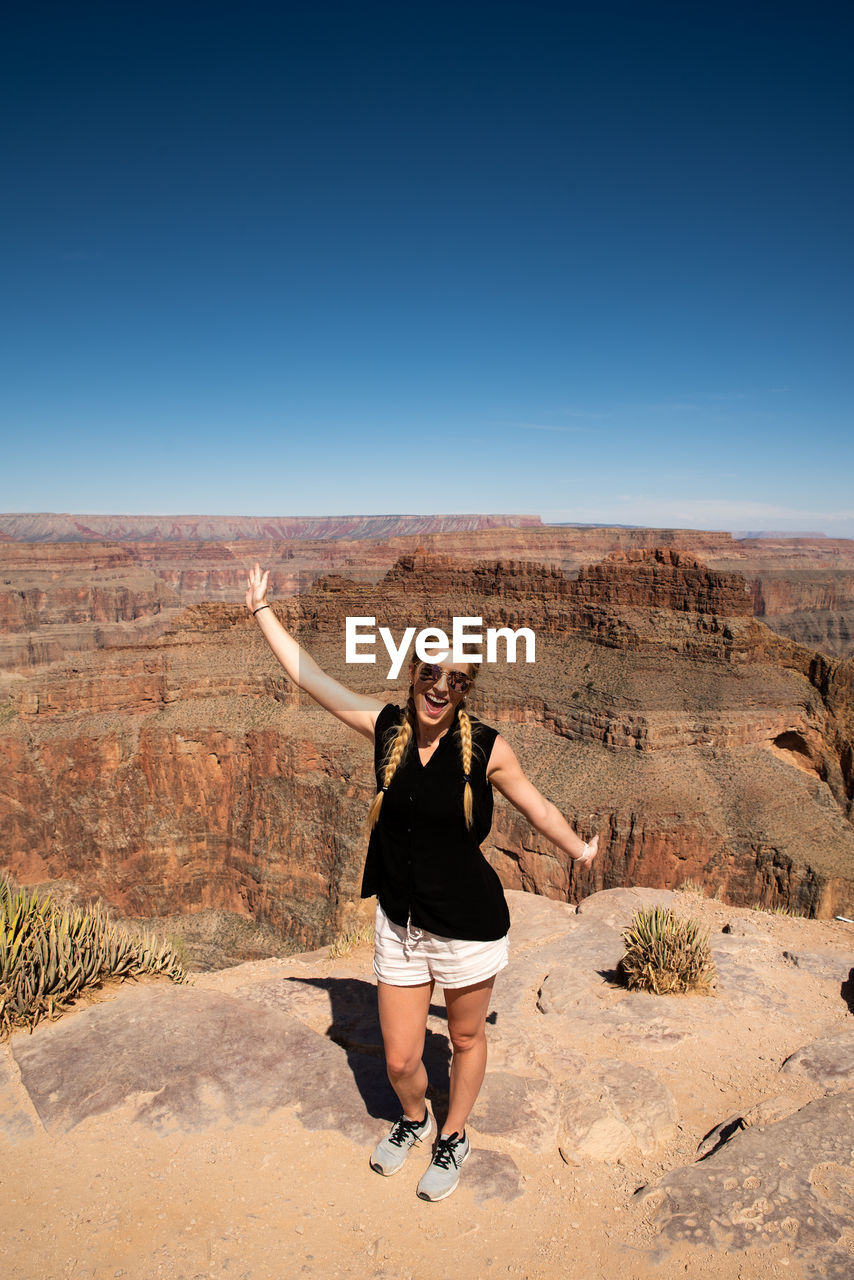 Portrait of young woman with arms raised standing on cliff against clear sky