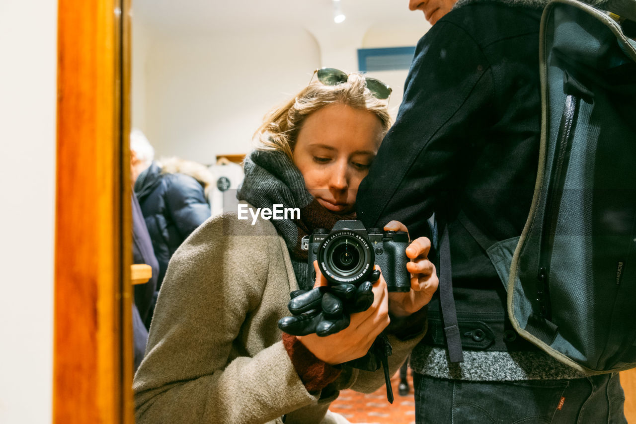 Woman holding camera while sitting by friend seen through glass door