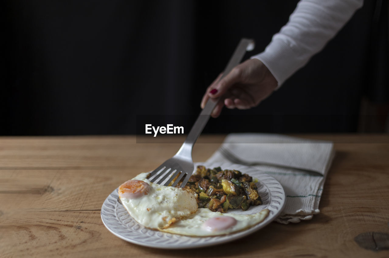 Female hand serves two fried eggs on a plate with stewed courgettes. black background.