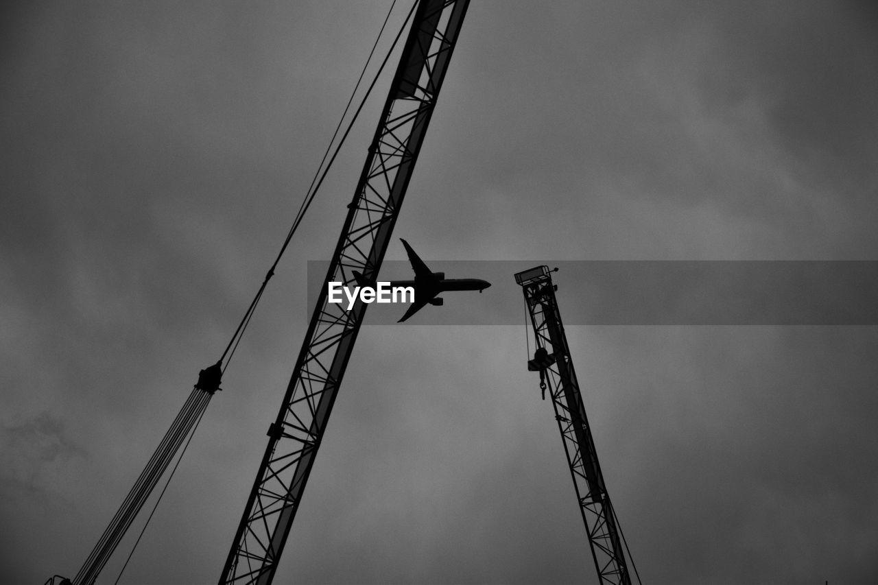 Low angle view of cranes and airplane against cloudy sky
