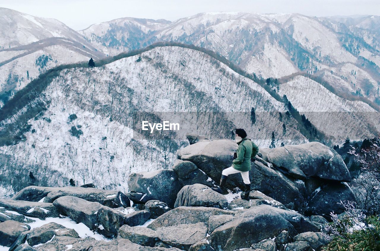 High angle view of man standing on rocks while looking at snowcapped mountains