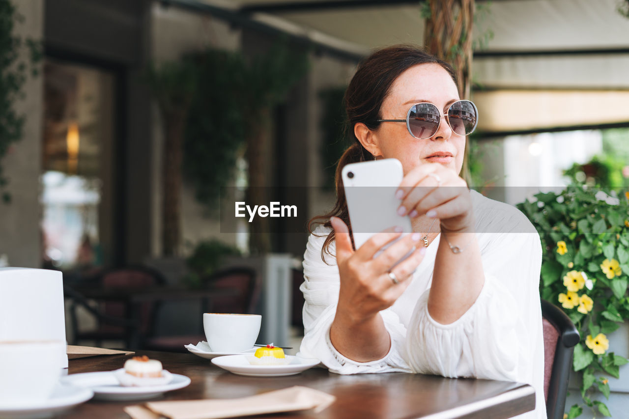 Beautiful middle aged woman in sunglasses in white clothes using mobile phone with coffee at cafe