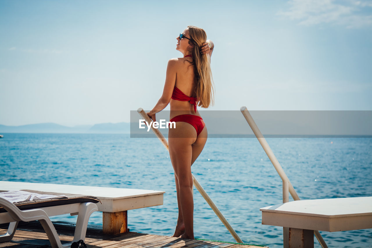 Young woman wearing red bikini while standing by sea against sky