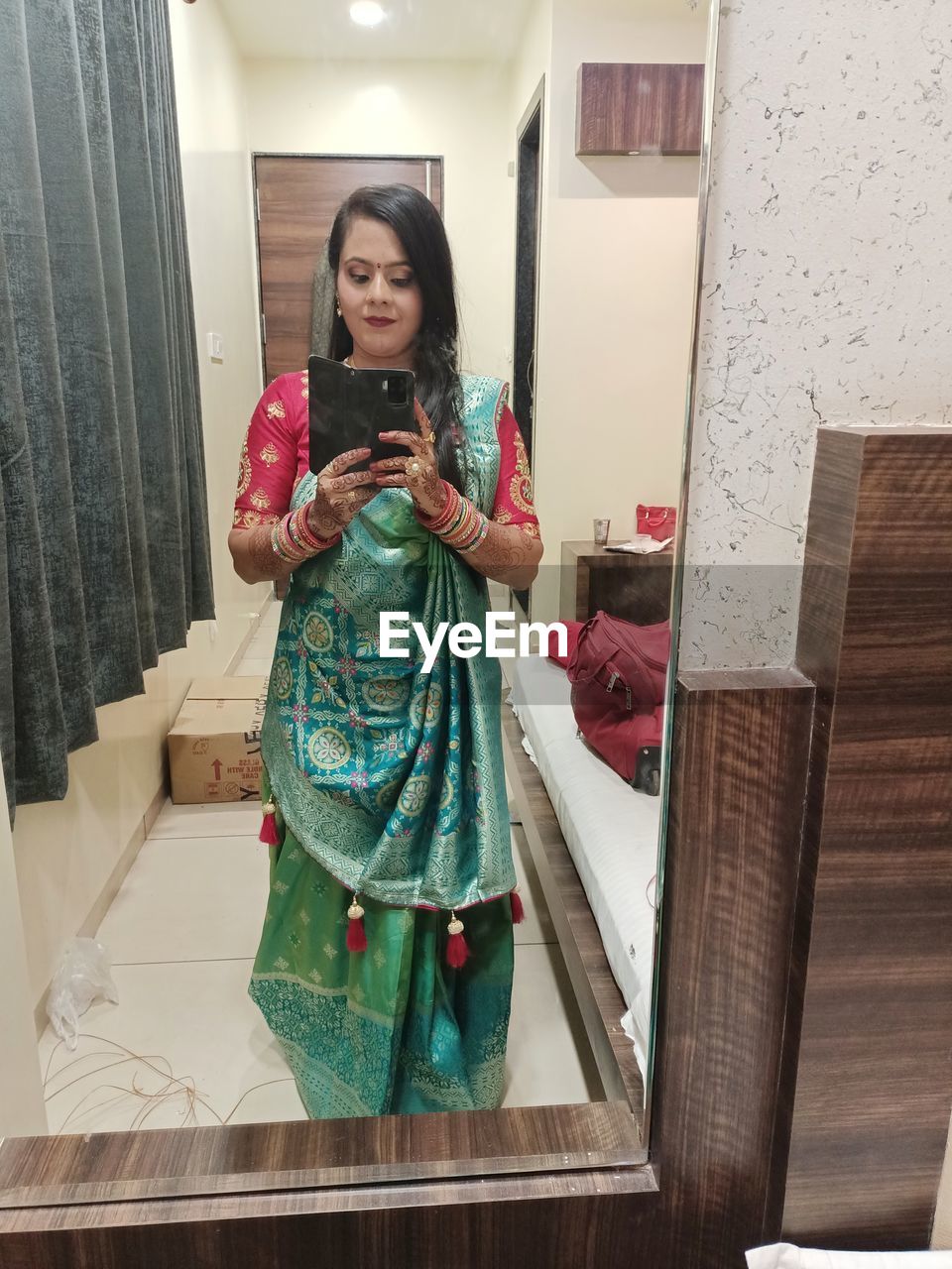 one person, women, adult, dress, indoors, clothing, smiling, front view, holding, standing, full length, happiness, lifestyles, young adult, mirror, portrait, female, technology, fashion, wireless technology, art, looking at camera, looking, communication, emotion, mobile phone, domestic room, person, architecture, traditional clothing, home interior