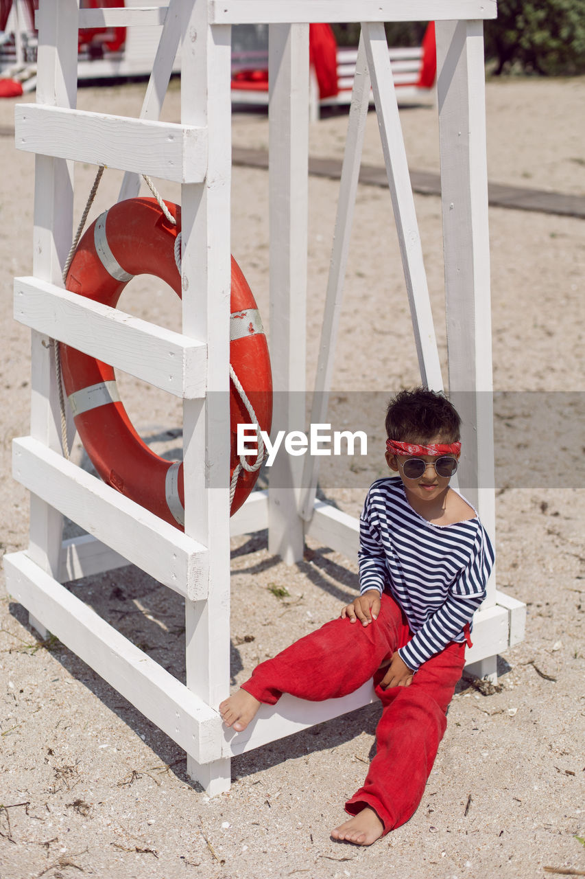 Child boy in striped clothes and red pants  standing on beach. white lifeguard tower, with a circle