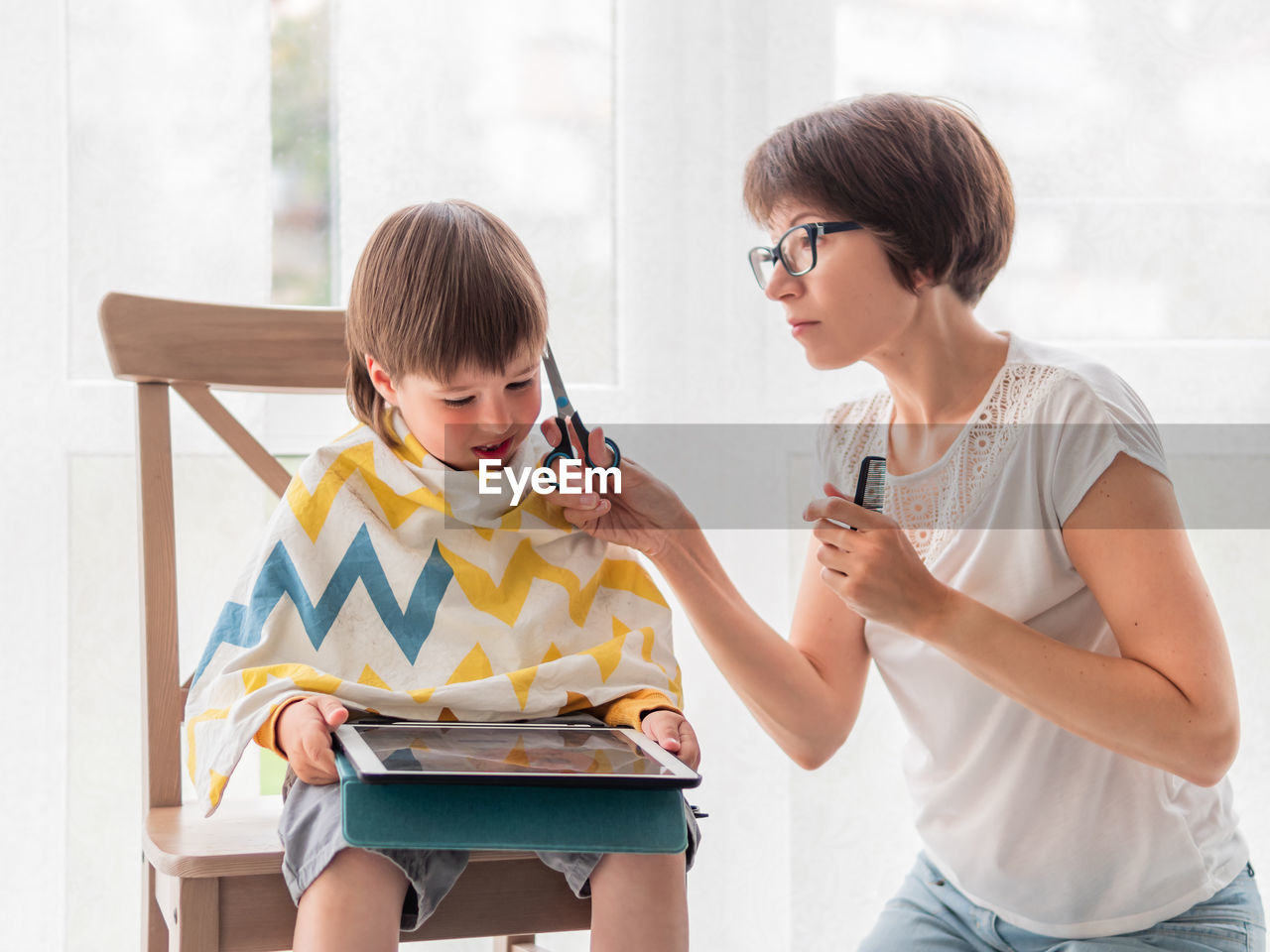 Mother cuts her son's hair by herself. little boy sits with digital tablet.