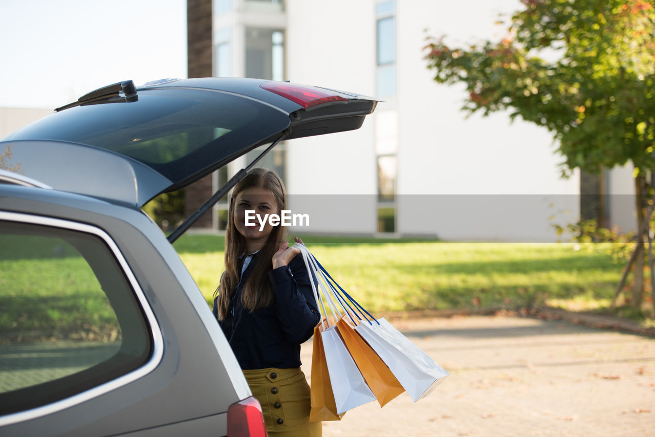 Portrait of smiling woman holding shopping bags while standing by car