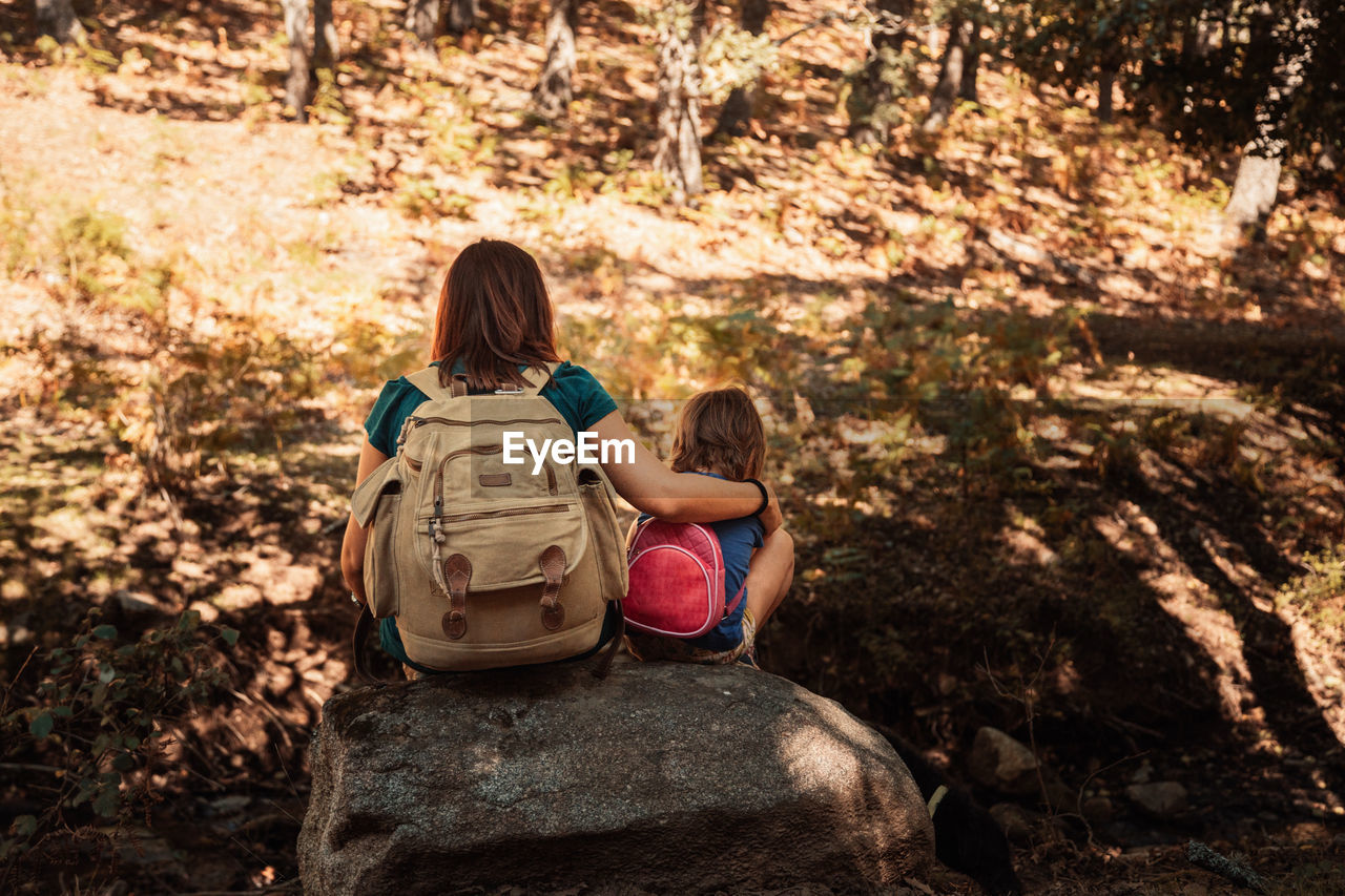 Rear view of mother and daughter with backpacks sitting on rocks in forest