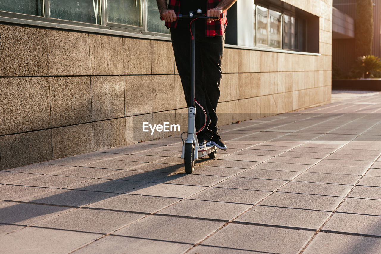 Unrecognizable crop male in checkered shirt riding modern electric scooter along pavement in city