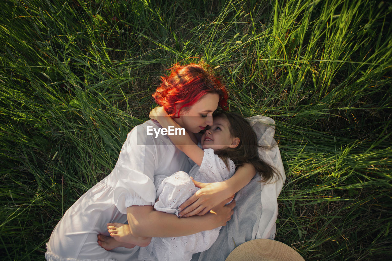 Girl a child hug mother mother with red hair lying on a green field at sunset