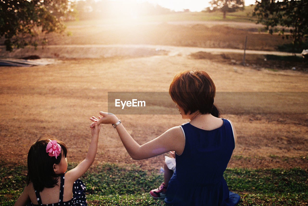 Rear view of mother and daughter holding hands while sitting on field during sunset