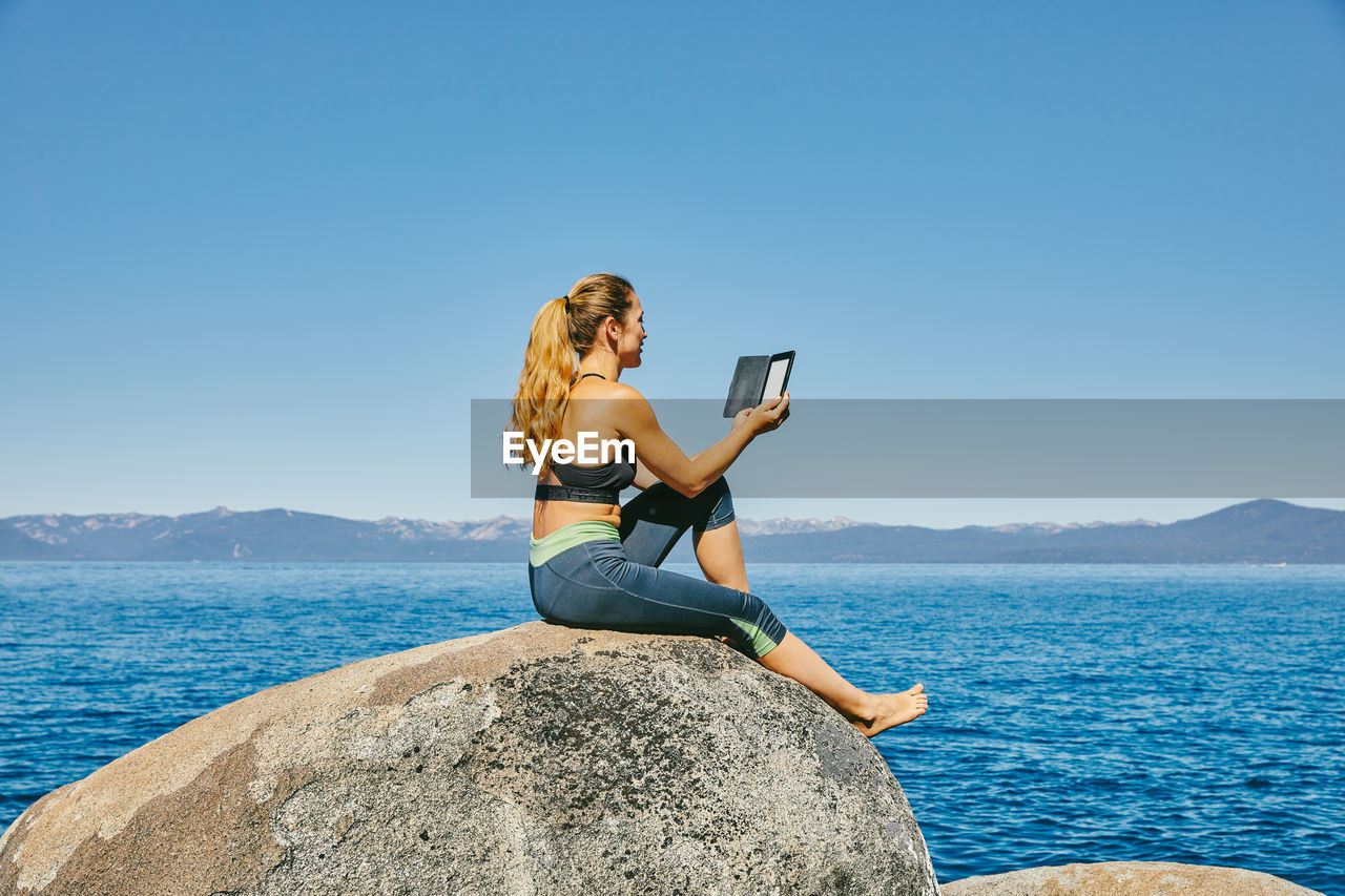 Young woman sitting by lake tahoe reading a kindle book during the day