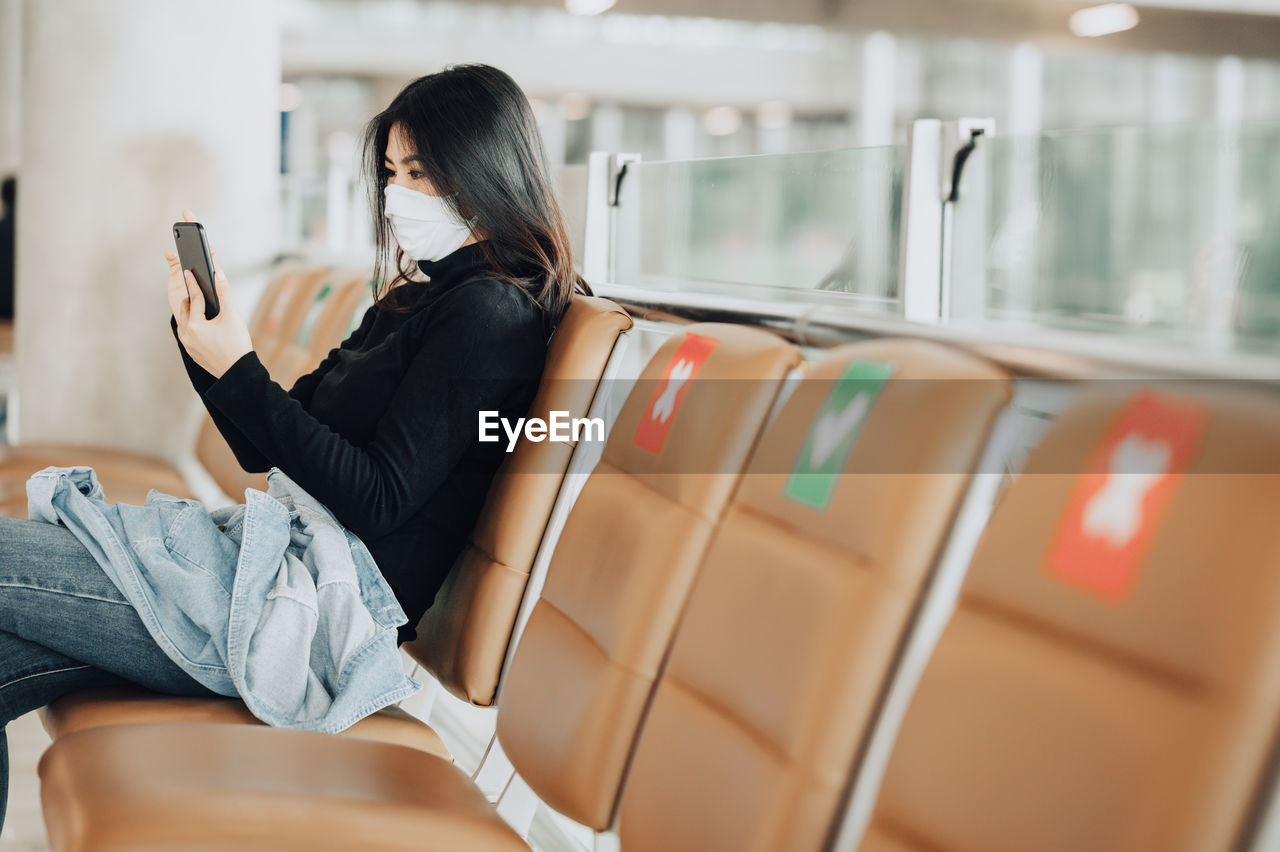 Woman wearing mask using smart phone sitting at airport