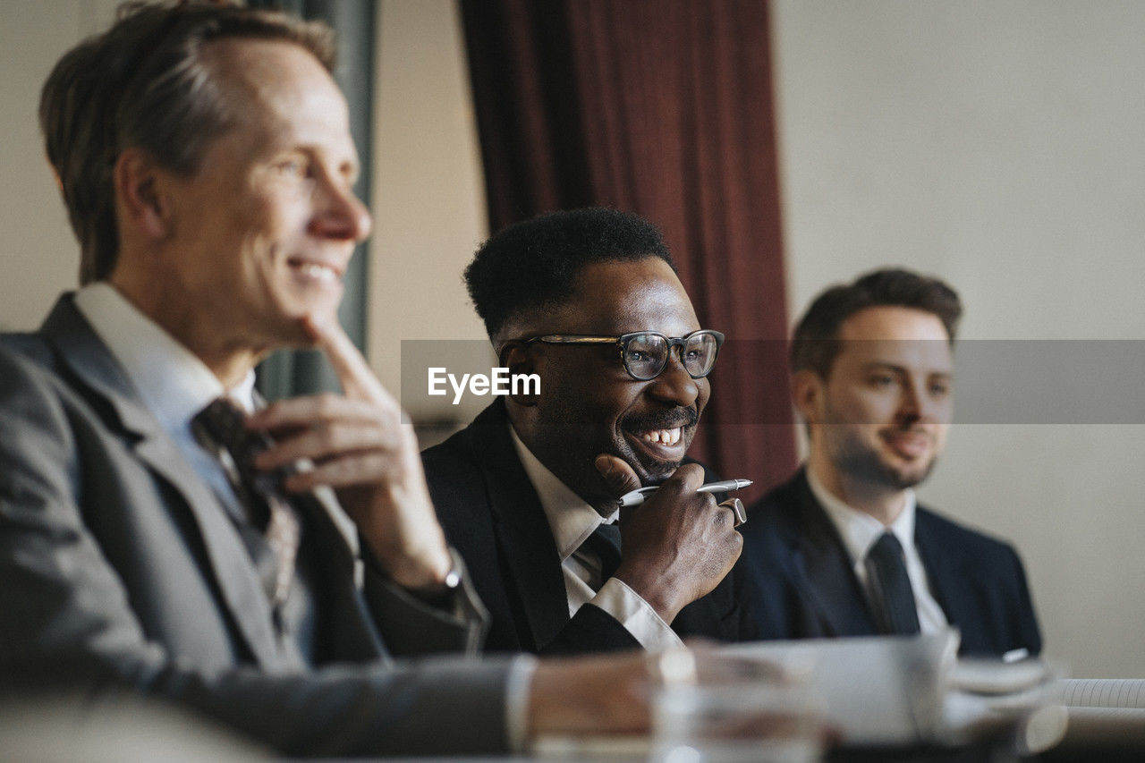 Smiling businessman with hand on chin sitting with male colleagues during meeting at office