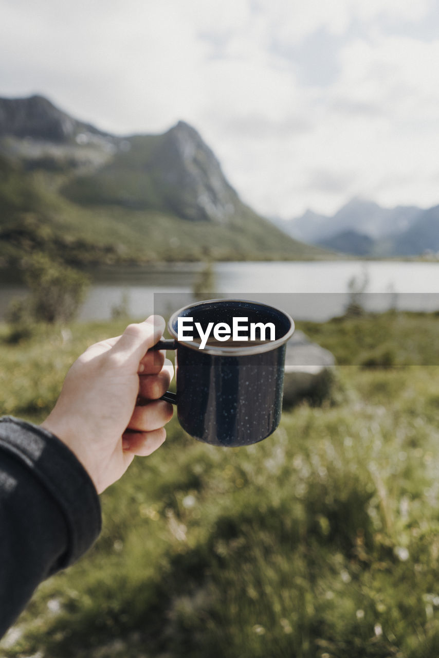 Cropped hand of person holding coffee cup against lake