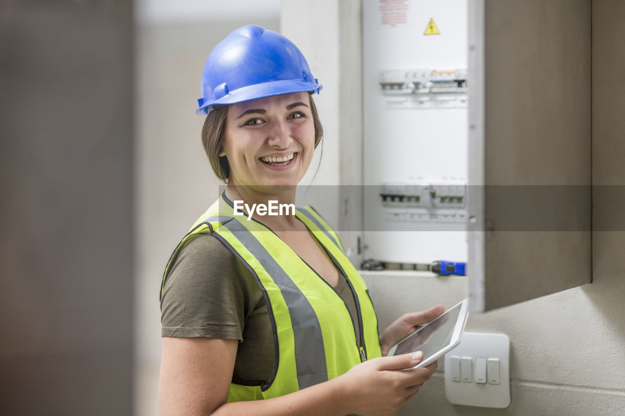 Portrait of smiling female electrician with tablet at fusebox