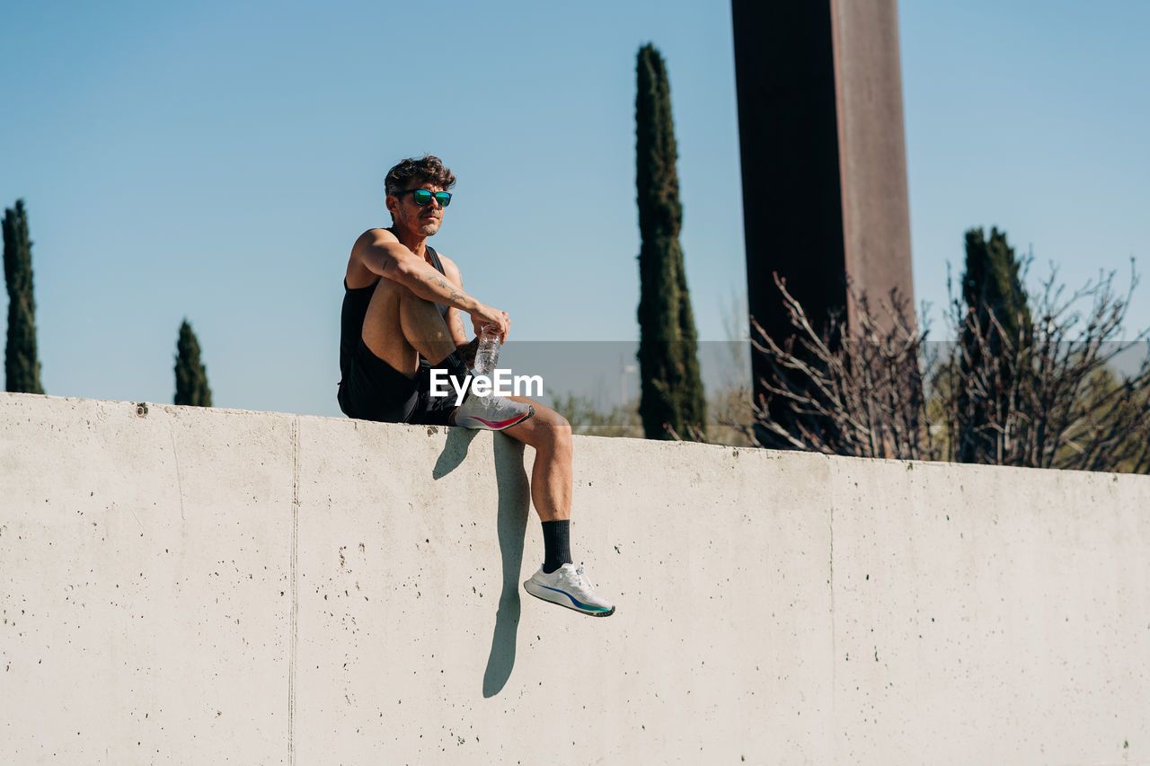 Sportsman in activewear with bottle of water after workout against trees in sunlight