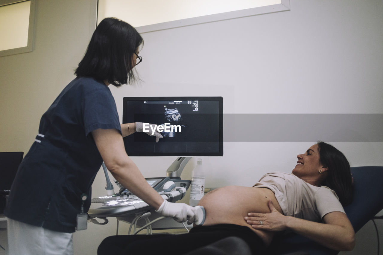 Female gynecologist pointing at screen while doing ultrasound of pregnant woman in medical clinic