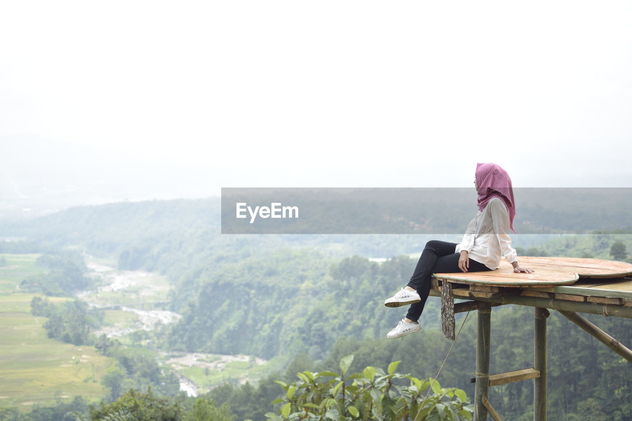 Woman looking at view while sitting on observation point against mountain and clear sky