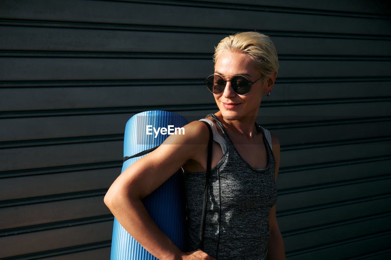 Sporty smiling blond woman in sportswear and sunglasses with yoga mat. workout outdoors. fitness