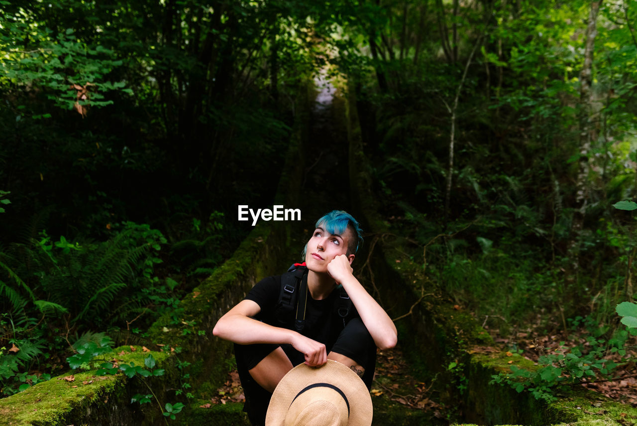 Relaxed traveling female with blue hair sitting in mossy woods during vacation and dreaming while looking up