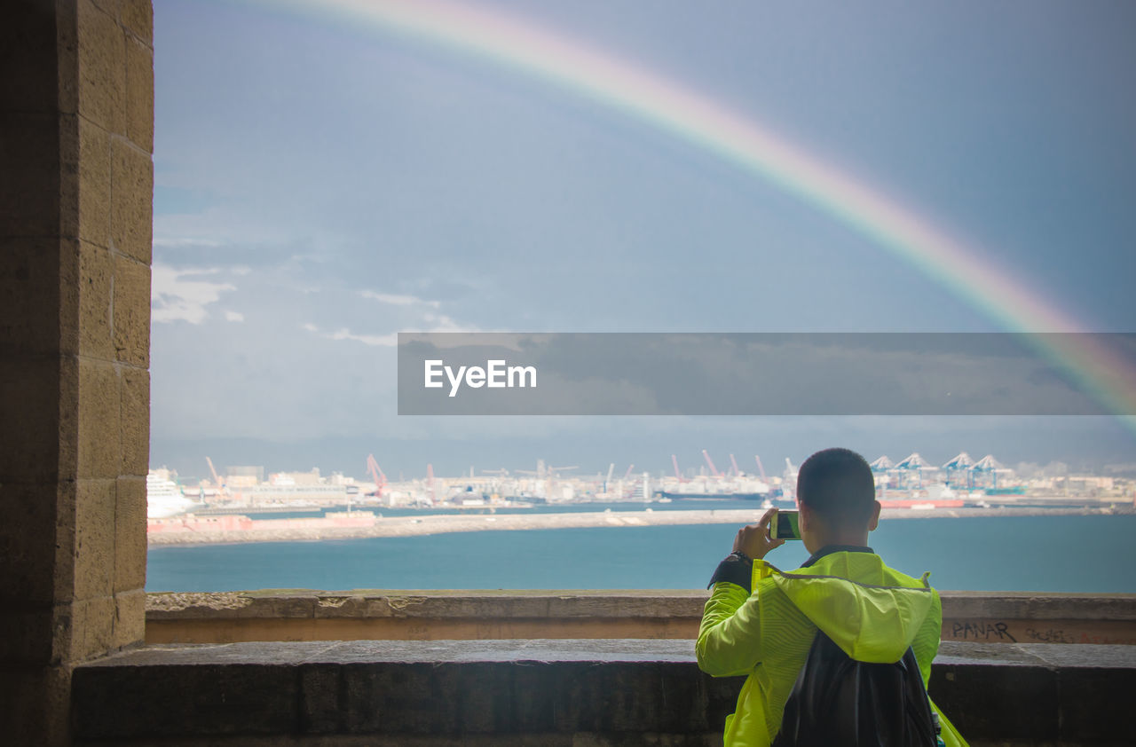 Rear view of man photographing standing by sea against rainbow in sky