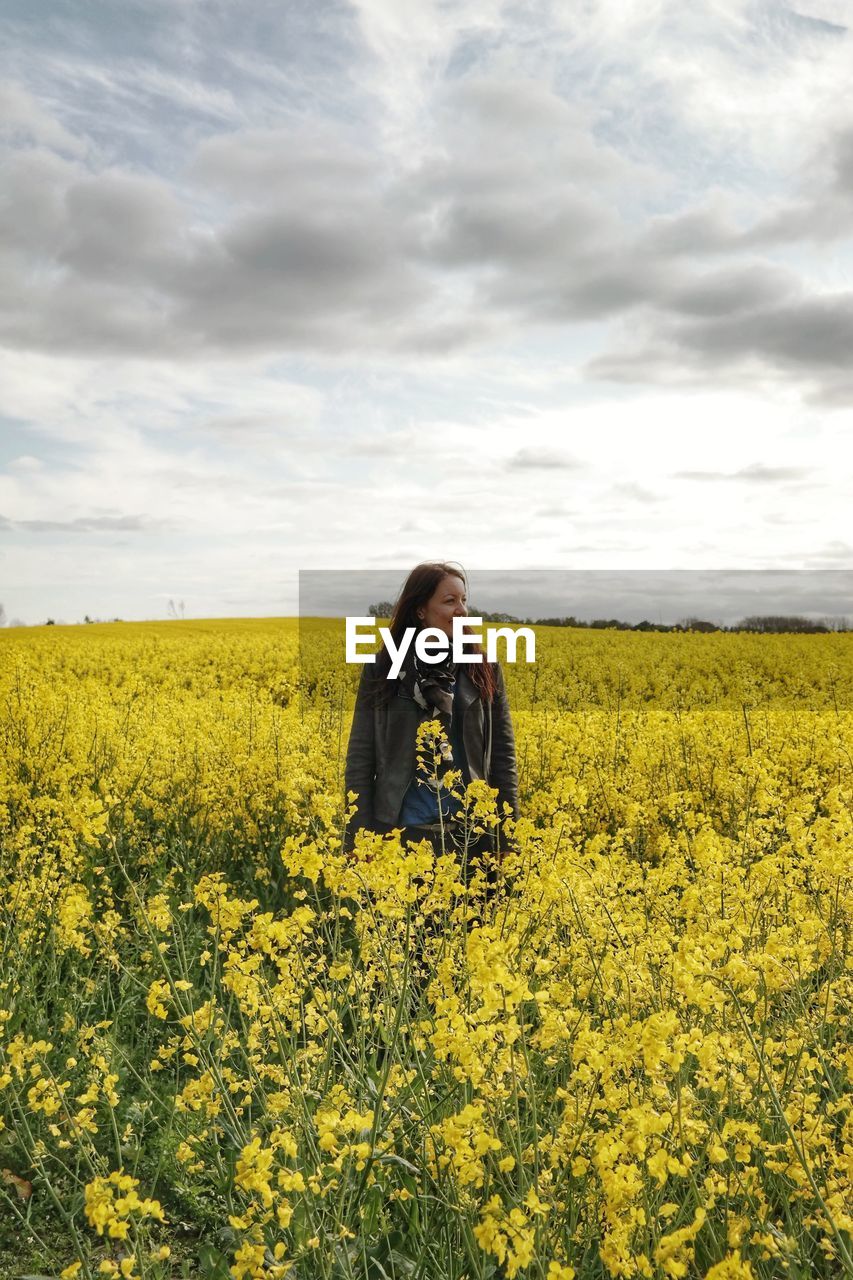 Woman standing in rapeseed field against cloudy sky