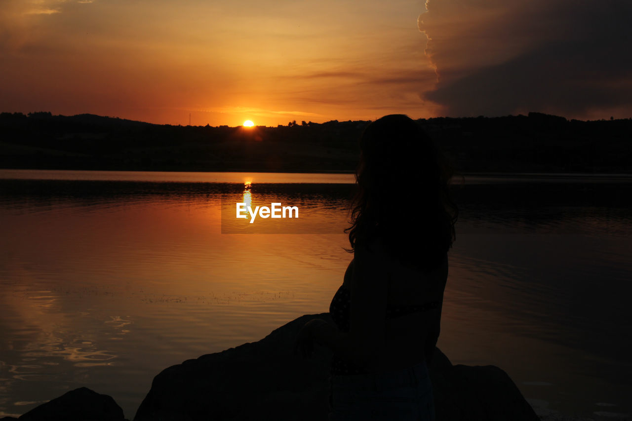 SILHOUETTE WOMAN BY LAKE AGAINST ORANGE SKY