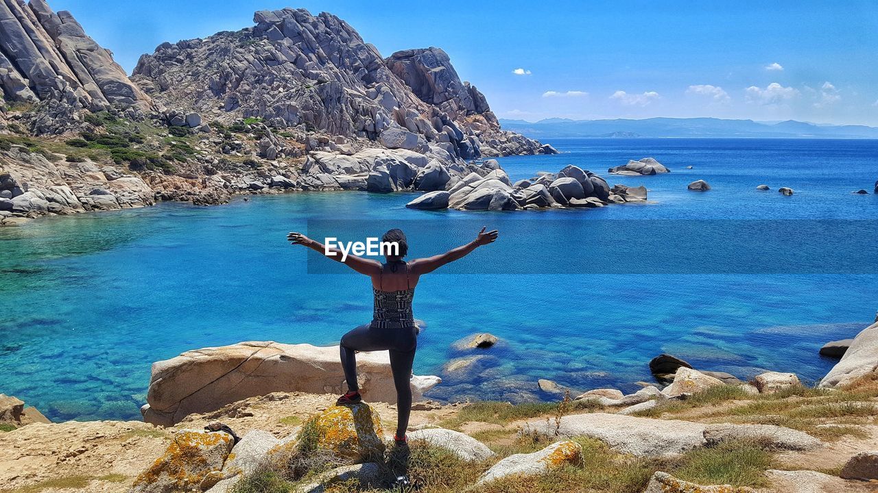 REAR VIEW OF MAN STANDING ON ROCK AGAINST SEA