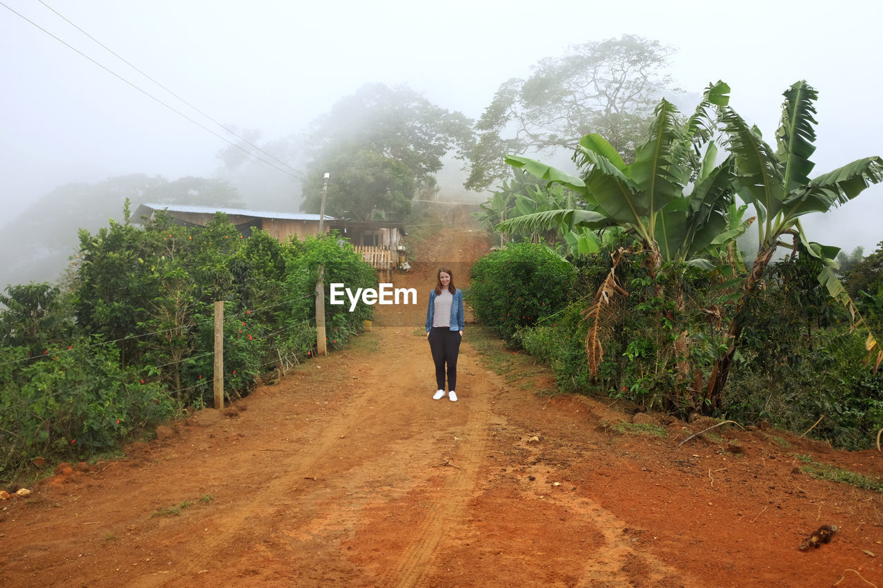 Full length of woman standing on dirt road amidst plants during foggy weather