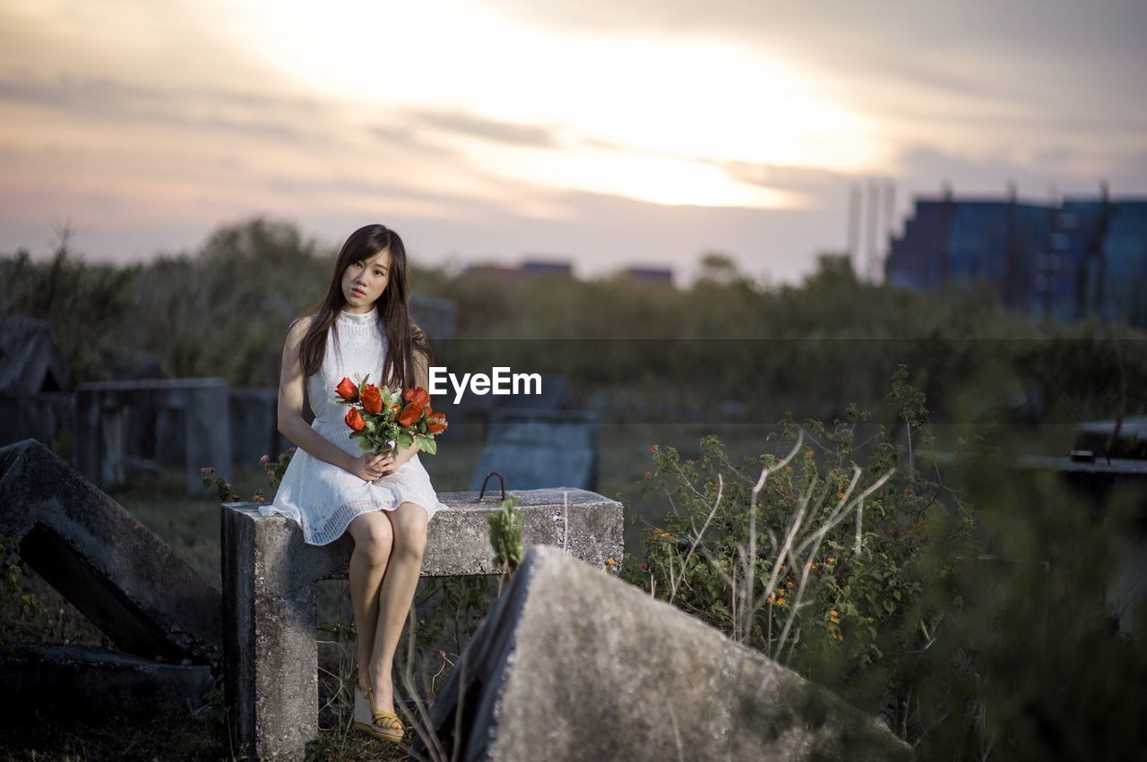 Portrait of young woman with flowers sitting on retaining wall against cloudy sky during sunset
