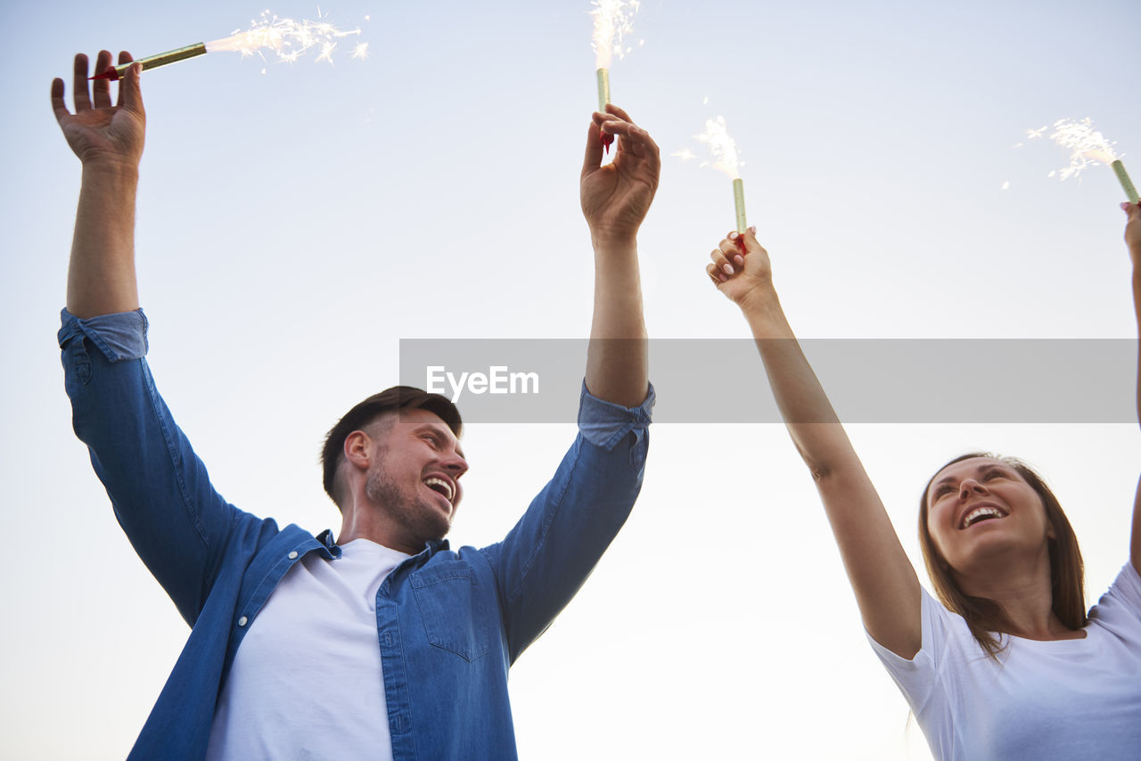 Low angle view of smiling couple holding lit sparklers against clear sky