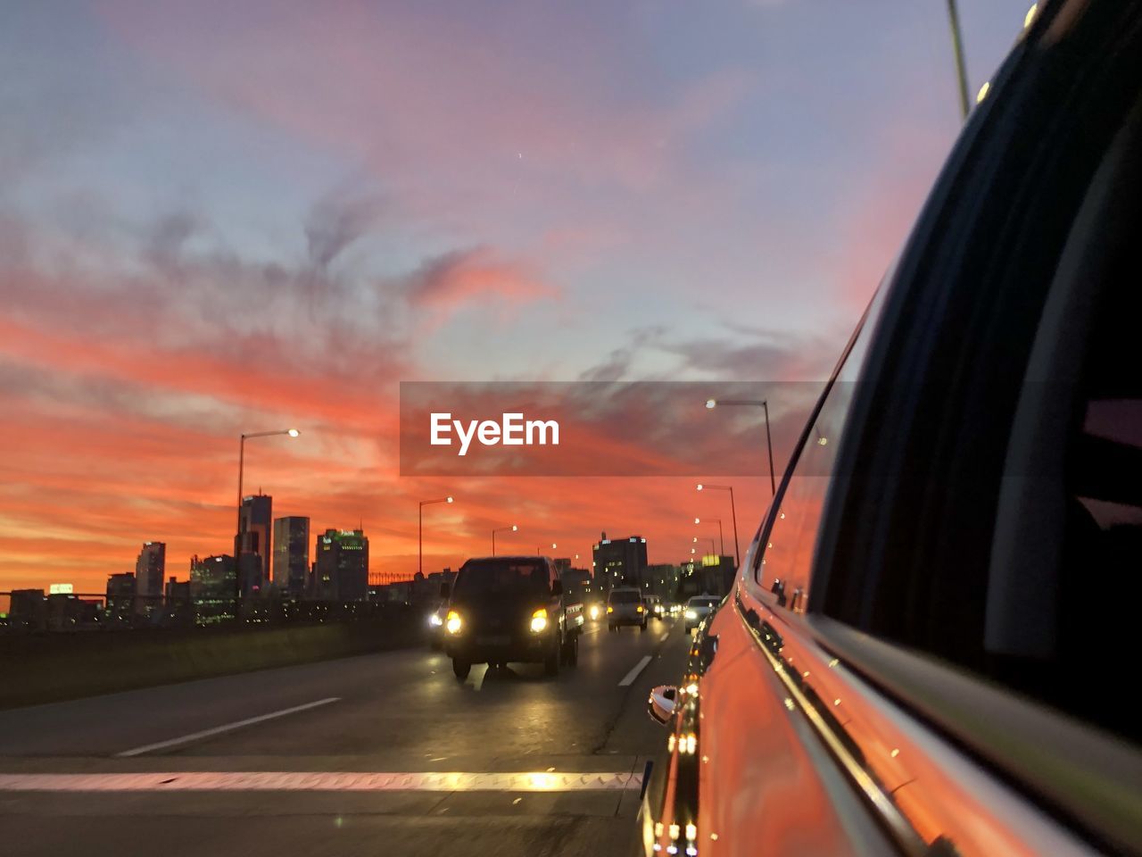 CARS ON ROAD AMIDST BUILDINGS AGAINST SKY DURING SUNSET