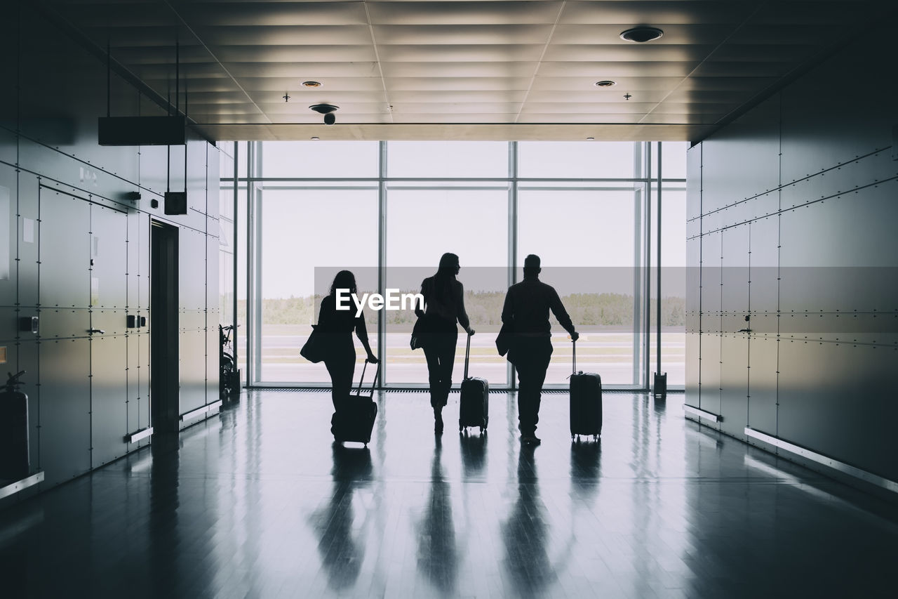 Rear view of silhouette business colleagues pulling luggage while walking in corridor at airport