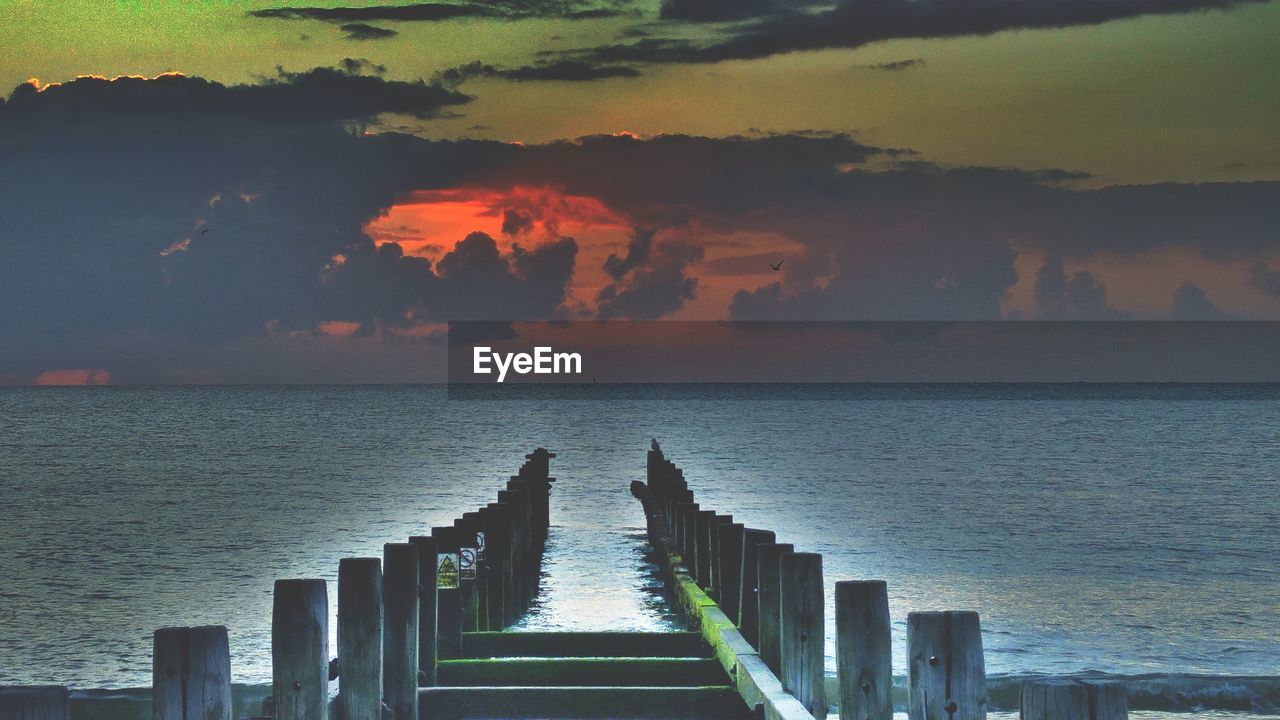 Wooden posts in sea against sky during sunset