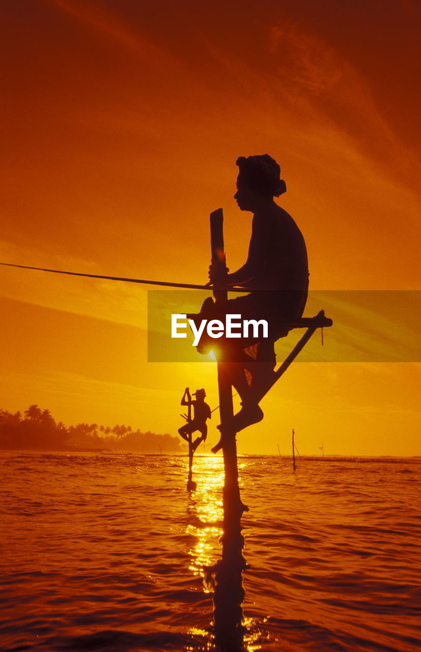 Low angle view of silhouette man stilt fishing in sea against orange sky