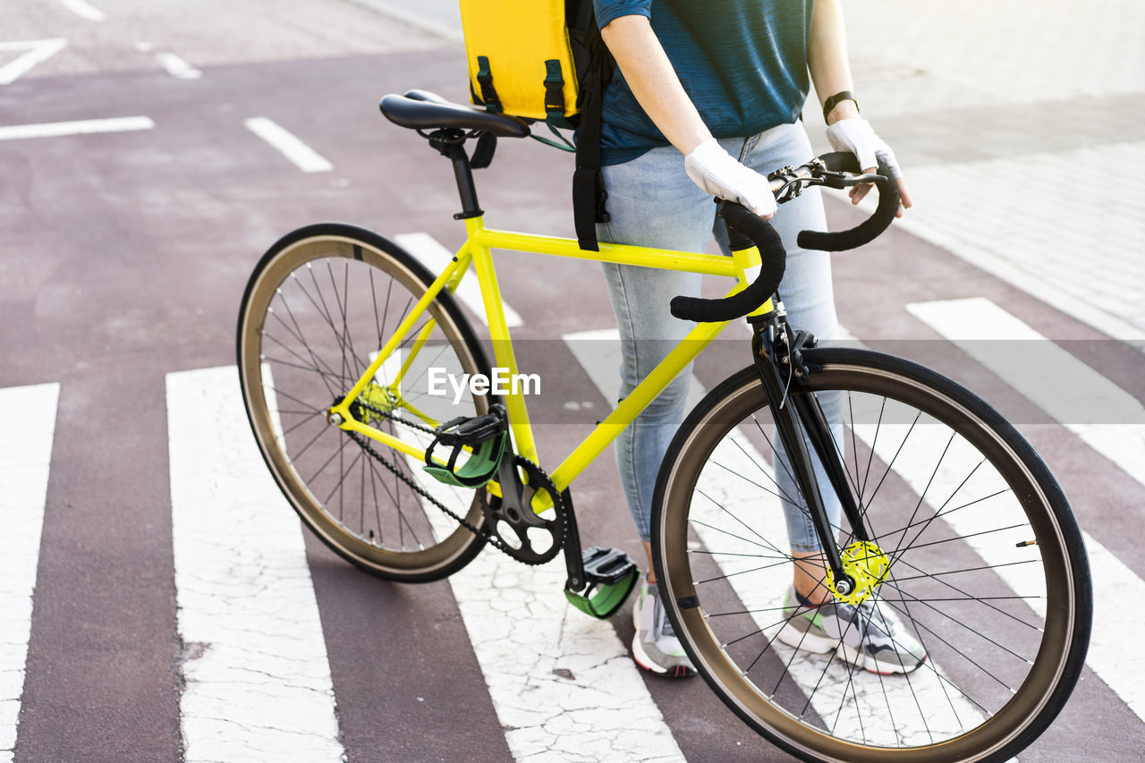 Young delivery woman with bicycle standing on zebra crossing