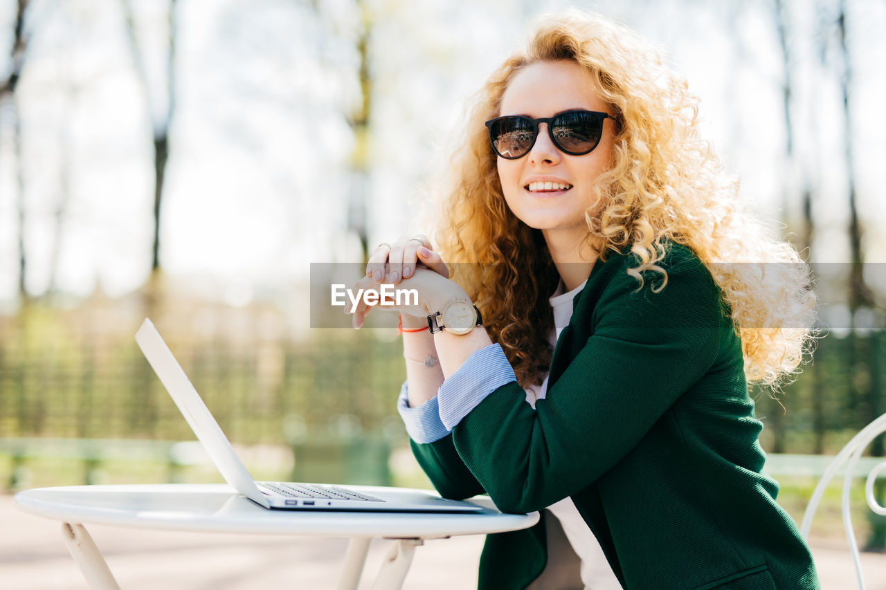 PORTRAIT OF YOUNG WOMAN USING MOBILE PHONE IN SUNGLASSES