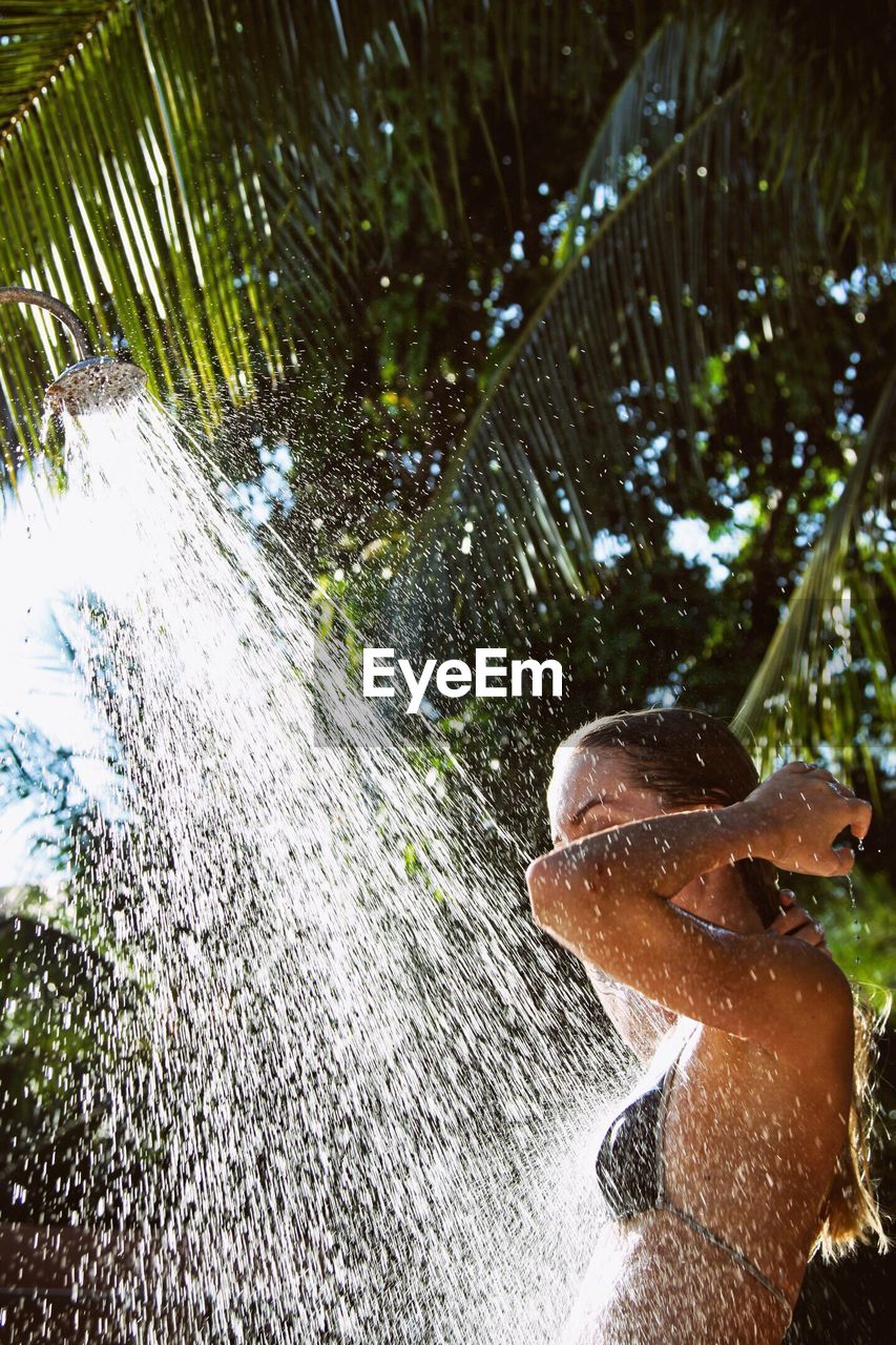 Side view of woman bathing under shower against trees