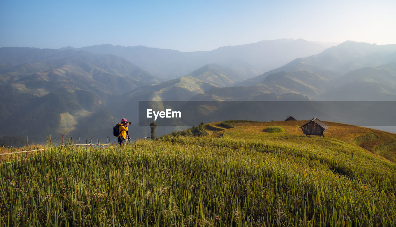 MAN STANDING ON RICE FIELD AGAINST MOUNTAINS