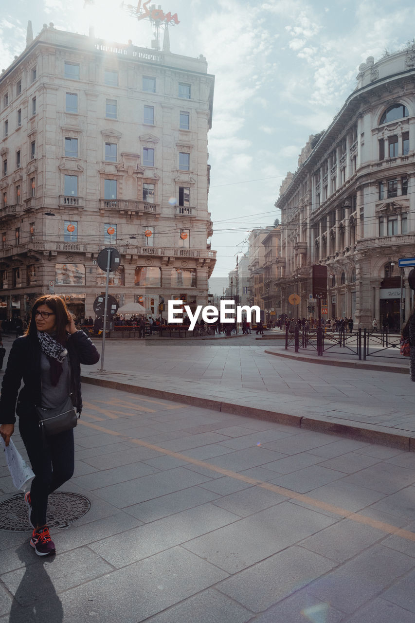 MAN PHOTOGRAPHING WOMAN STANDING ON STREET AGAINST BUILDINGS