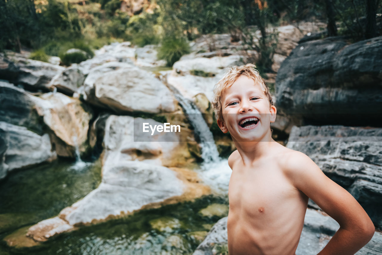 Portrait of happy shirtless boy standing by stream in forest