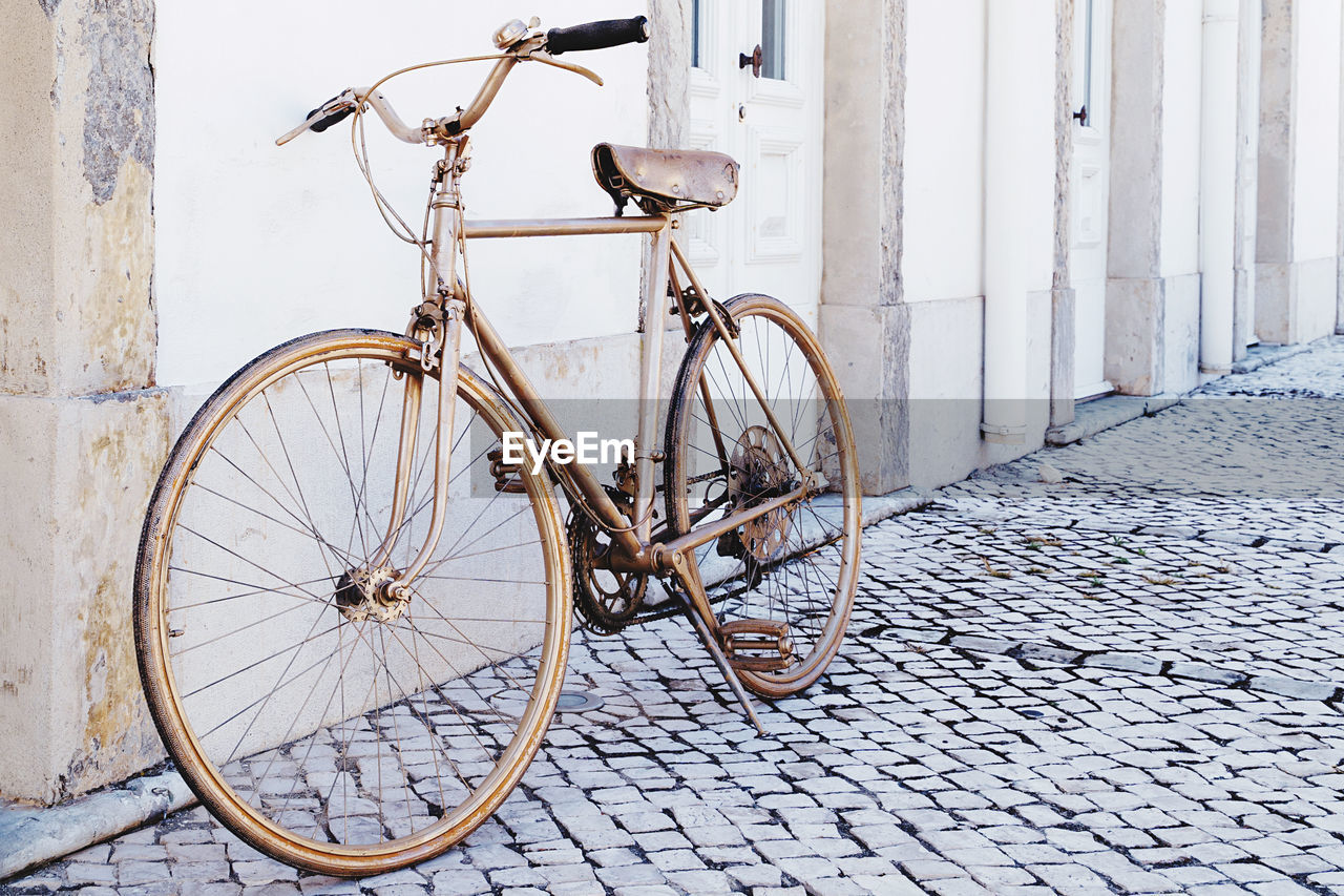 bicycle, transportation, architecture, mode of transportation, vehicle, city, road bicycle, land vehicle, street, no people, sports equipment, building exterior, wall - building feature, built structure, wheel, day, footpath, old, outdoors, travel, sidewalk, cobblestone, building, leaning, city life, white, nature, parking, iron, bicycle wheel, city street