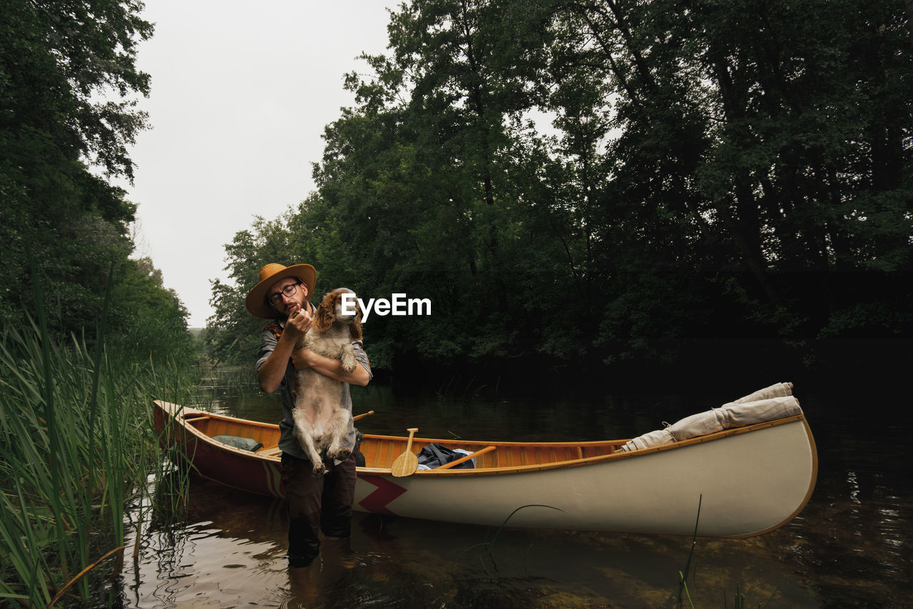 Young man stands in shallow river next to his canoe with a dog. adventurous active rest concept